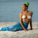 Person dressed as a mermaid sitting on sandy beach with a blue mermaid tail and shell bikini top, evoking a touch of boudoir photography.