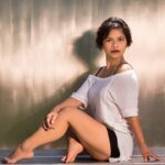 Person seated on the floor, wearing a white top and black shorts, with a neutral expression. Background is a blurred, metallic texture that subtly whispers,